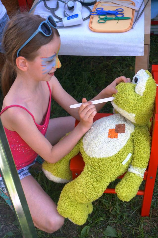 Dziewczynka z pomalowaną twarzą próbuje otworzyć szpatułką lekarską buzię maskotce-żabie podczas festynu Czerwcówka pod Kopcem (Powstania Warszawskiego) na Mokotowie