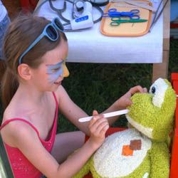 Miniatura: Dziewczynka z pomalowaną twarzą próbuje otworzyć szpatułką lekarską buzię maskotce-żabie podczas festynu Czerwcówka pod Kopcem Powstania Warszawskiego na Mokotowie