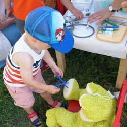 Miniatura: Chłopczyk udający wyciąganie kleszcza z nogi pluszaka-żaby na stoisku przychodni pediatrycznej Eskulapek na Mokotowie podczas festynu